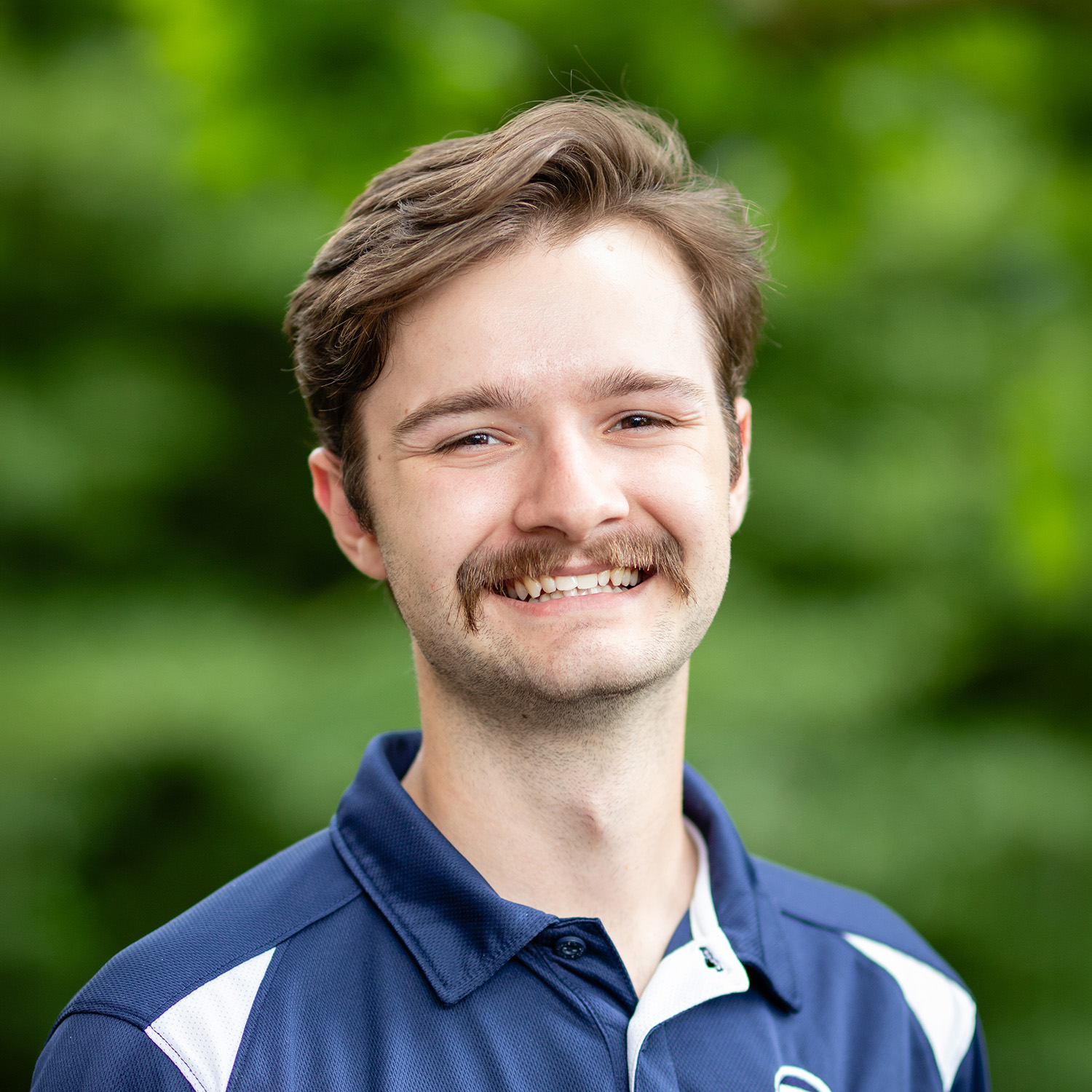 Headshot of Ryan Predki, a man with brown hair and navy polo shirt smiling against a background of a flowering tree