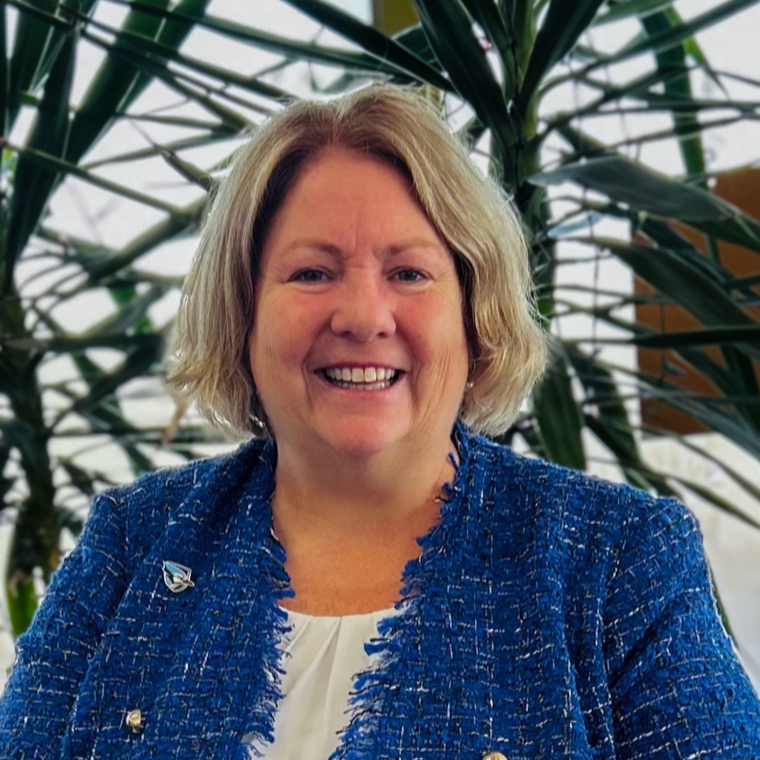 Headshot of Missy Gillis, a woman with short sandy blonde hair and navy tweed jacket with a white blouse smiling against a background of foliage